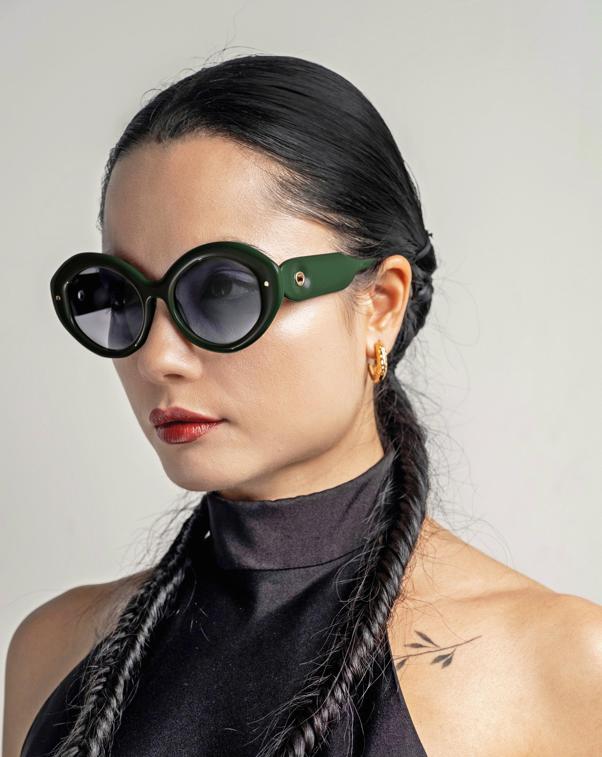 A woman with long, black hair styled in two braids and wearing large, round Helios eyewear by For Art&#39;s Sake®. She has gold hoop earrings with gold plating and a tattoo of an arrow on her shoulder. She is dressed in a sleeveless black top against a plain background.
