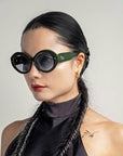 A woman with long, black hair styled in two braids and wearing large, round Helios eyewear by For Art's Sake®. She has gold hoop earrings with gold plating and a tattoo of an arrow on her shoulder. She is dressed in a sleeveless black top against a plain background.