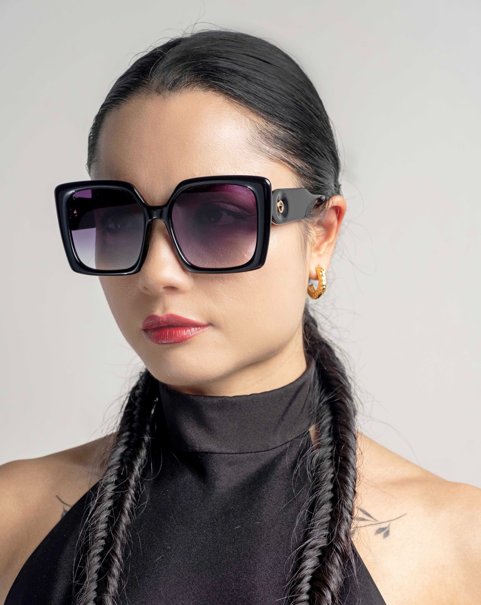 A person with long, dark braided hair is wearing large, square-shaped sunglasses with classic temple round details and a black halter-top. They have a pair of hoop earrings and an 18 karat gold plating accent, all while sporting a contemplative expression, looking slightly to the side against a plain, light-colored background. They are also wearing Eos by For Art&#39;s Sake®.