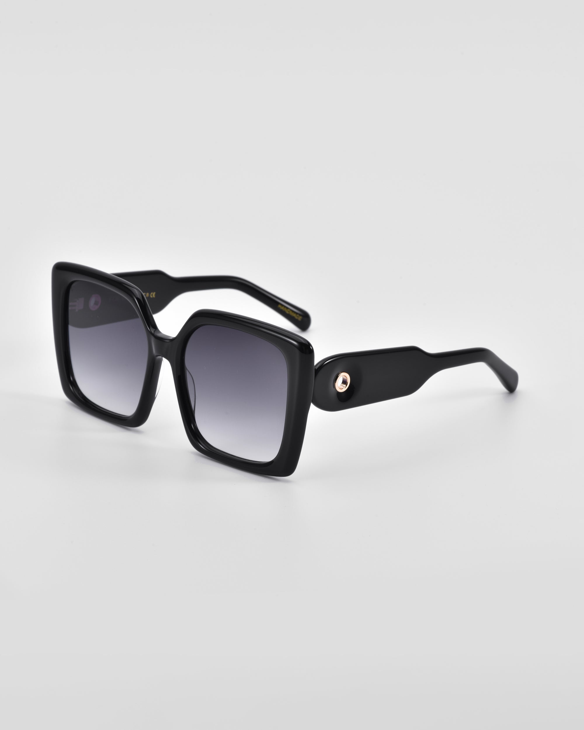A pair of black oversized soft-square silhouette sunglasses with gradient dark lenses rests on a light grey surface. The arms of the sunglasses are thick and feature classic temple round details near the hinge. The overall design is sleek and modern. These are the Eos by For Art&#39;s Sake®.