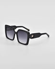 A pair of black oversized soft-square silhouette sunglasses with gradient dark lenses rests on a light grey surface. The arms of the sunglasses are thick and feature classic temple round details near the hinge. The overall design is sleek and modern. These are the Eos by For Art's Sake®.