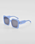 A pair of oversized soft-square Eos sunglasses from For Art's Sake® with thick, light blue frames and dark gray lenses. The earpieces feature classic temple round details, are slightly curved, and the overall design is modern and stylish. The background is plain white.