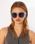 A woman with light brown hair wears oversized soft-square Eos sunglasses with classic temple round details by For Art's Sake® and has a neutral facial expression. She is dressed in a gray sleeveless top and stands against a plain white background.