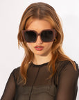 A woman with shoulder-length, light brown hair is wearing elegant eyewear featuring an oversized soft-square silhouette from For Art's Sake® Eos and a black, sheer long-sleeve top. She is standing against a plain white background.