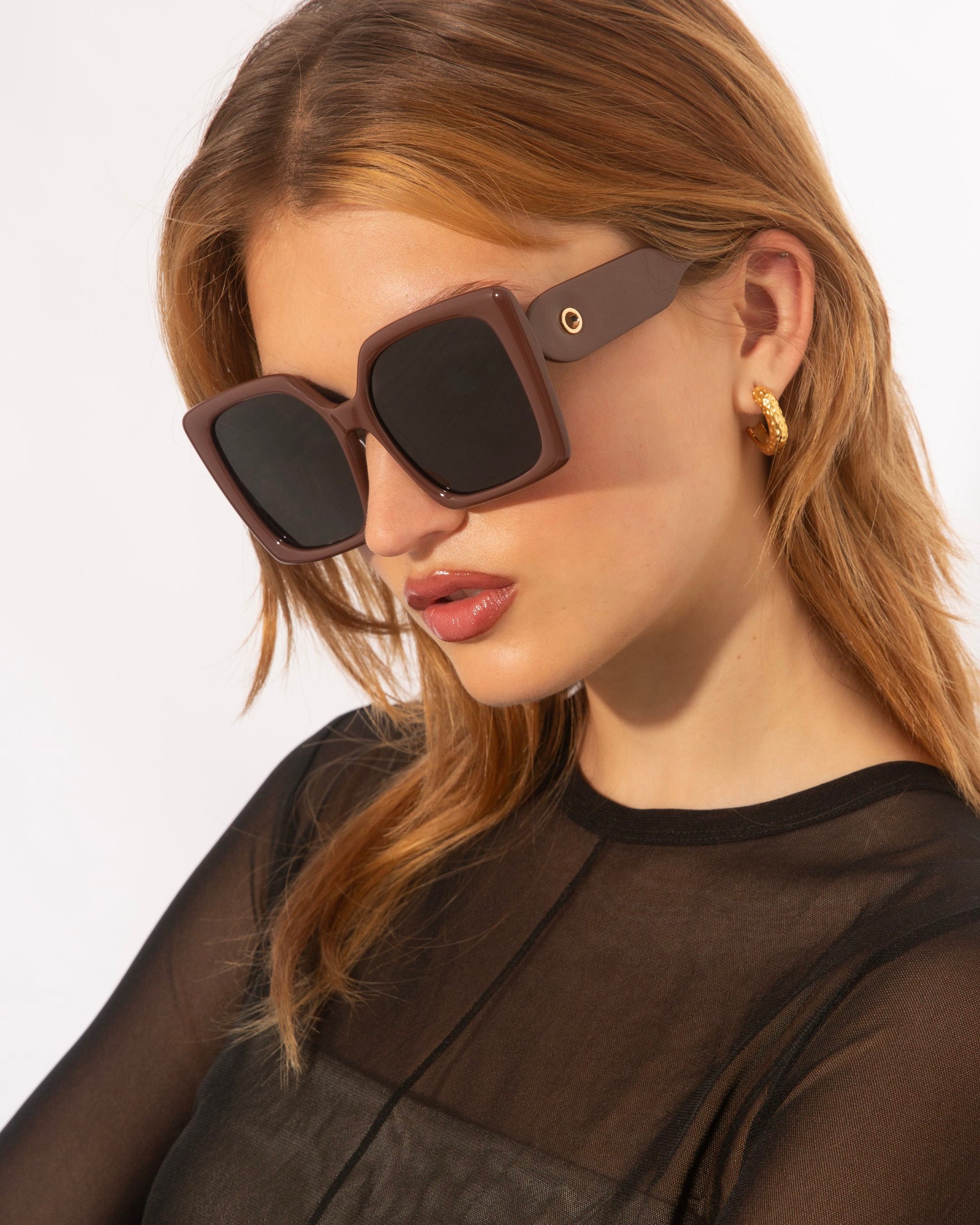 A person with shoulder-length light brown hair, wearing elegant eyewear characterized by oversized soft-square silhouettes and a pair of gold hoop earrings, is looking downward. The person is dressed in a sheer black top with a high neckline. The background is plain white. They are wearing Eos by For Art's Sake®.