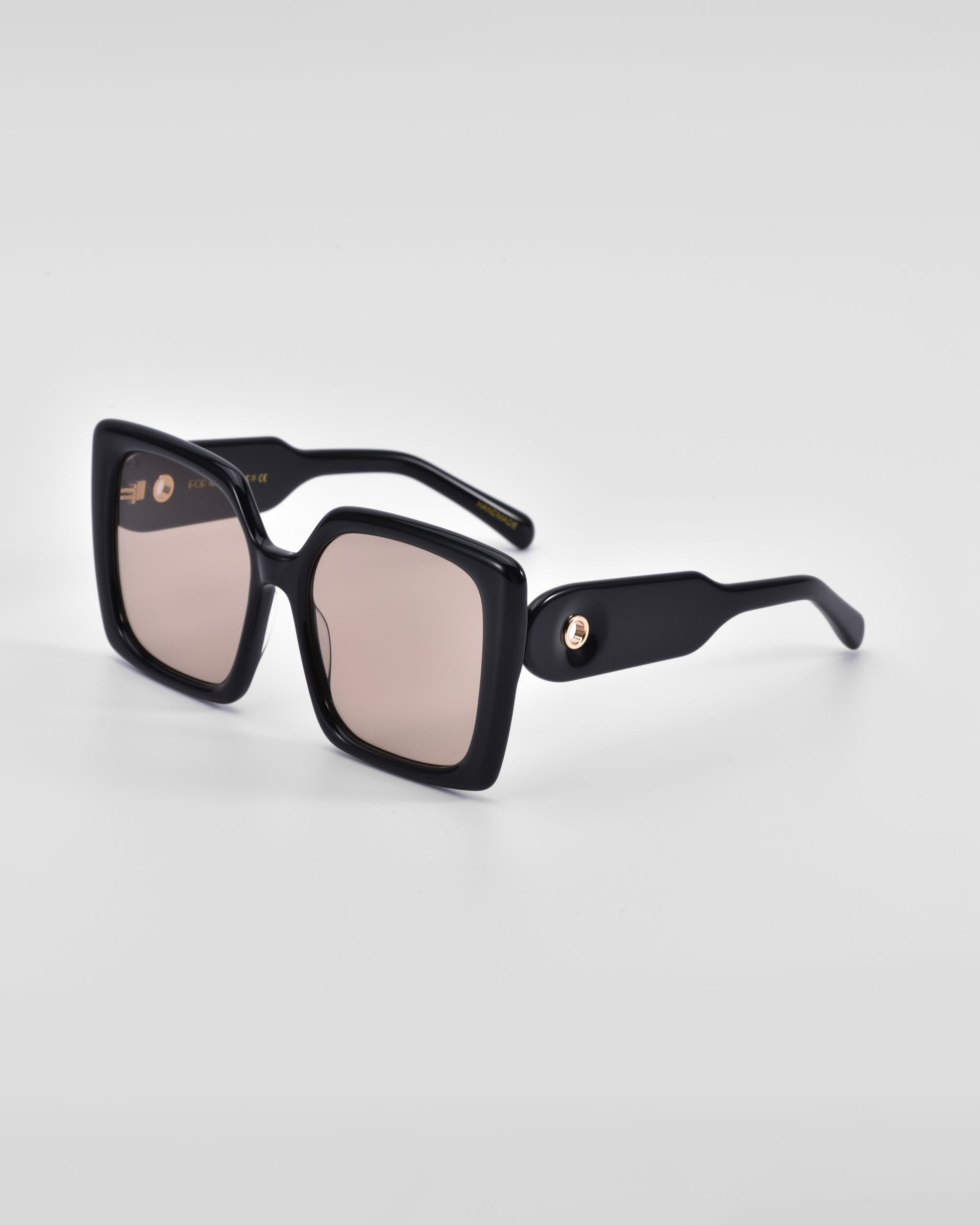 A pair of For Art&#39;s Sake® Eos sunglasses with rectangular black frames and light brown tinted lenses is displayed against a light gray background. The temples feature an 18 karat gold circular ornament near the hinges.