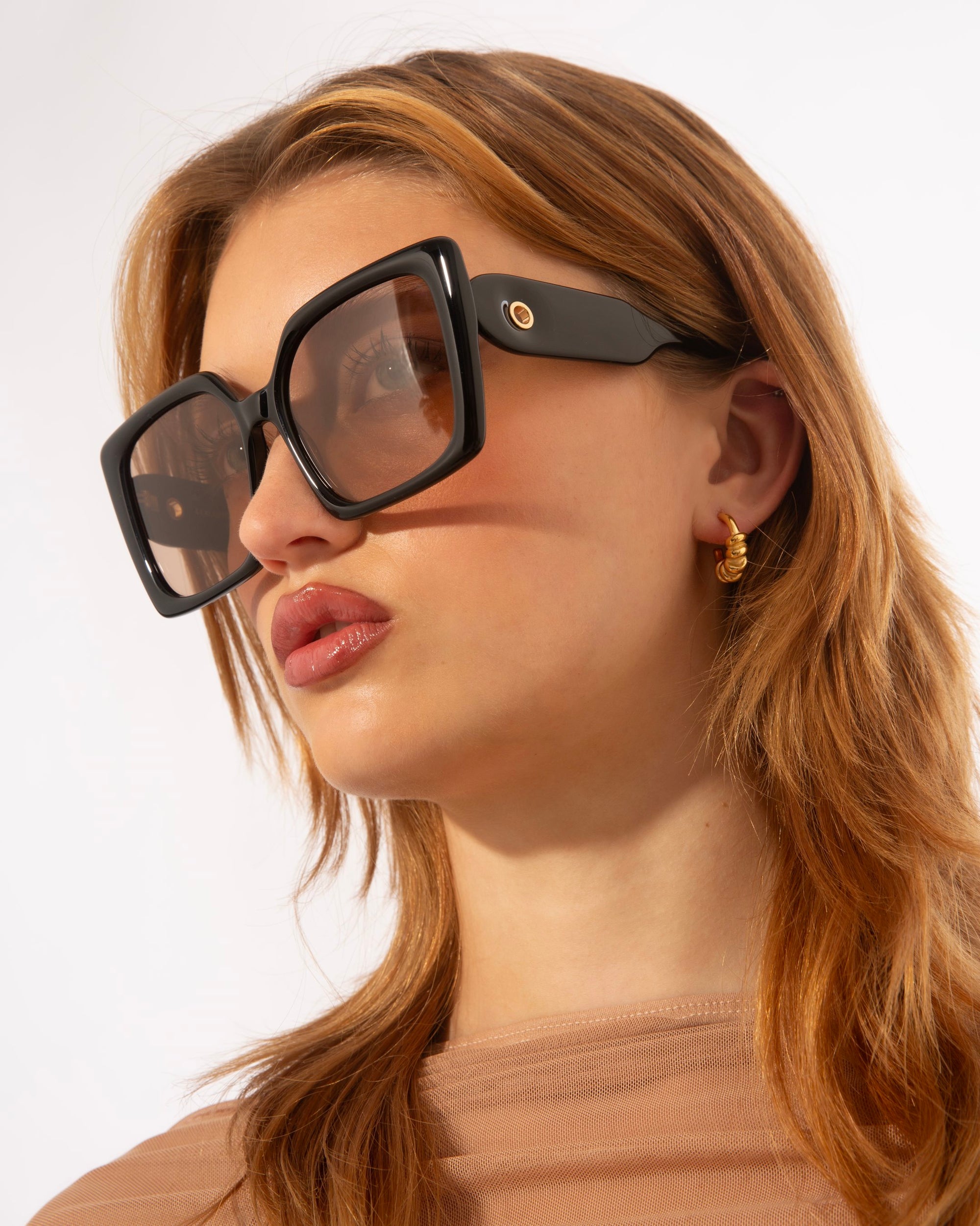A person with reddish-brown hair wears oversized square Eos sunglasses by For Art&#39;s Sake® and gold hoop earrings. They are dressed in a light brown top. The background is a clean, light color, highlighting the stylish accessories and hair.