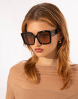 A person with light brown hair is wearing Eos by For Art's Sake® oversized soft-square sunglasses with classic temple round details and 18 karat gold plating. They also sport gold hoop earrings and a sheer, nude-colored top, looking off to the side against a plain white background.