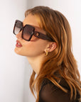 A woman with long, wavy auburn hair is wearing oversized Eos soft-square silhouette sunglasses by For Art's Sake® with dark frames and lenses. She has small gold hoop earrings featuring 18 karat gold plating and is looking upwards slightly. The background is plain and white.