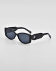 A pair of black rectangular Mene sunglasses from For Art's Sake® with dark lenses and thick arms, featuring a small circular metallic detail on the hinges. The background is a plain, light gray surface. These luxury frames exude sophistication and timeless style.