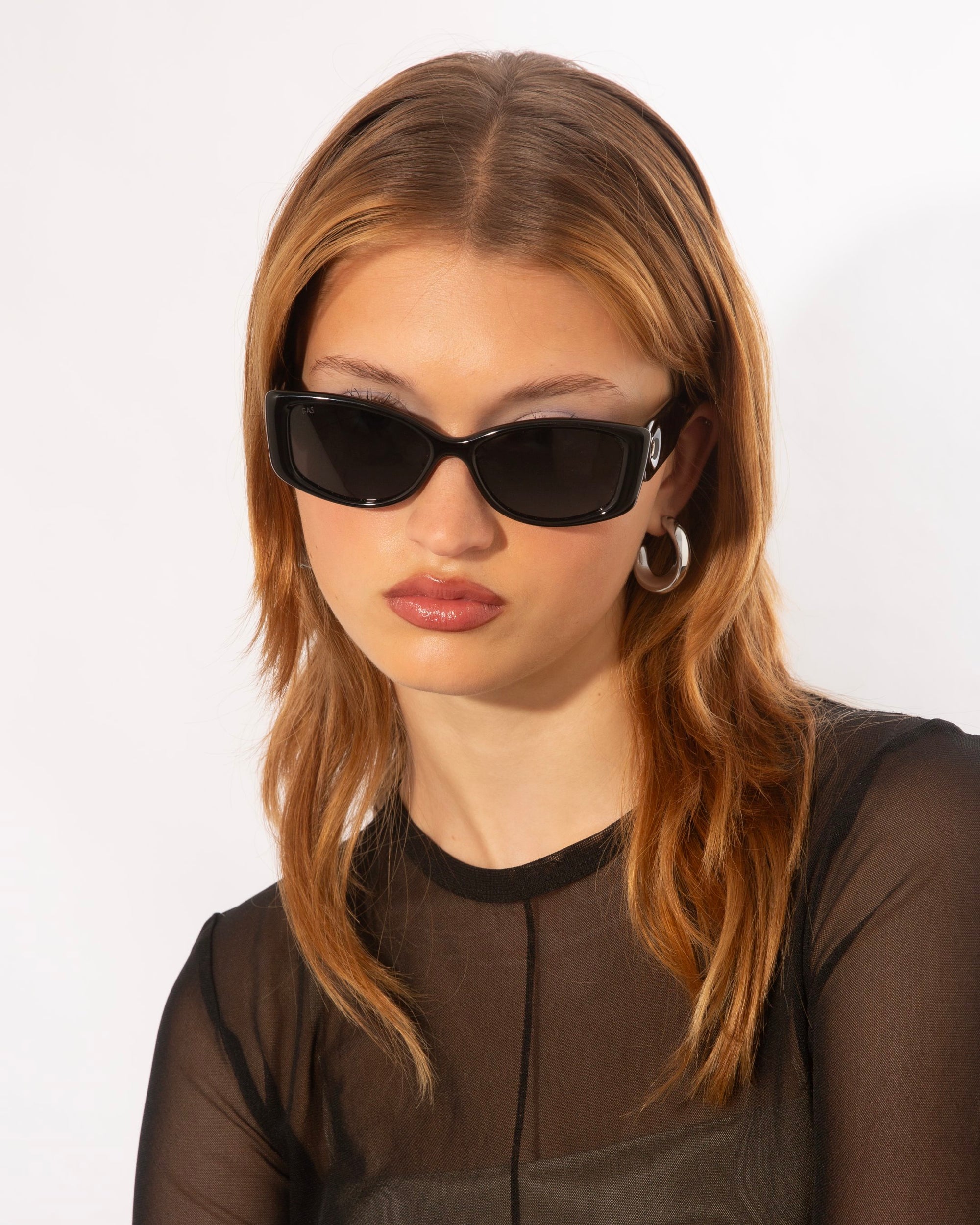 A person with light brown hair, wearing rectangular black For Art's Sake® Mene sunglasses and silver hoop earrings. Clad in a sheer black top, they have a calm expression and their head slightly tilted, embodying luxury eyewear style against a plain white background.