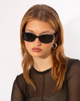 A person with light brown hair, wearing rectangular black For Art's Sake® Mene sunglasses and silver hoop earrings. Clad in a sheer black top, they have a calm expression and their head slightly tilted, embodying luxury eyewear style against a plain white background.