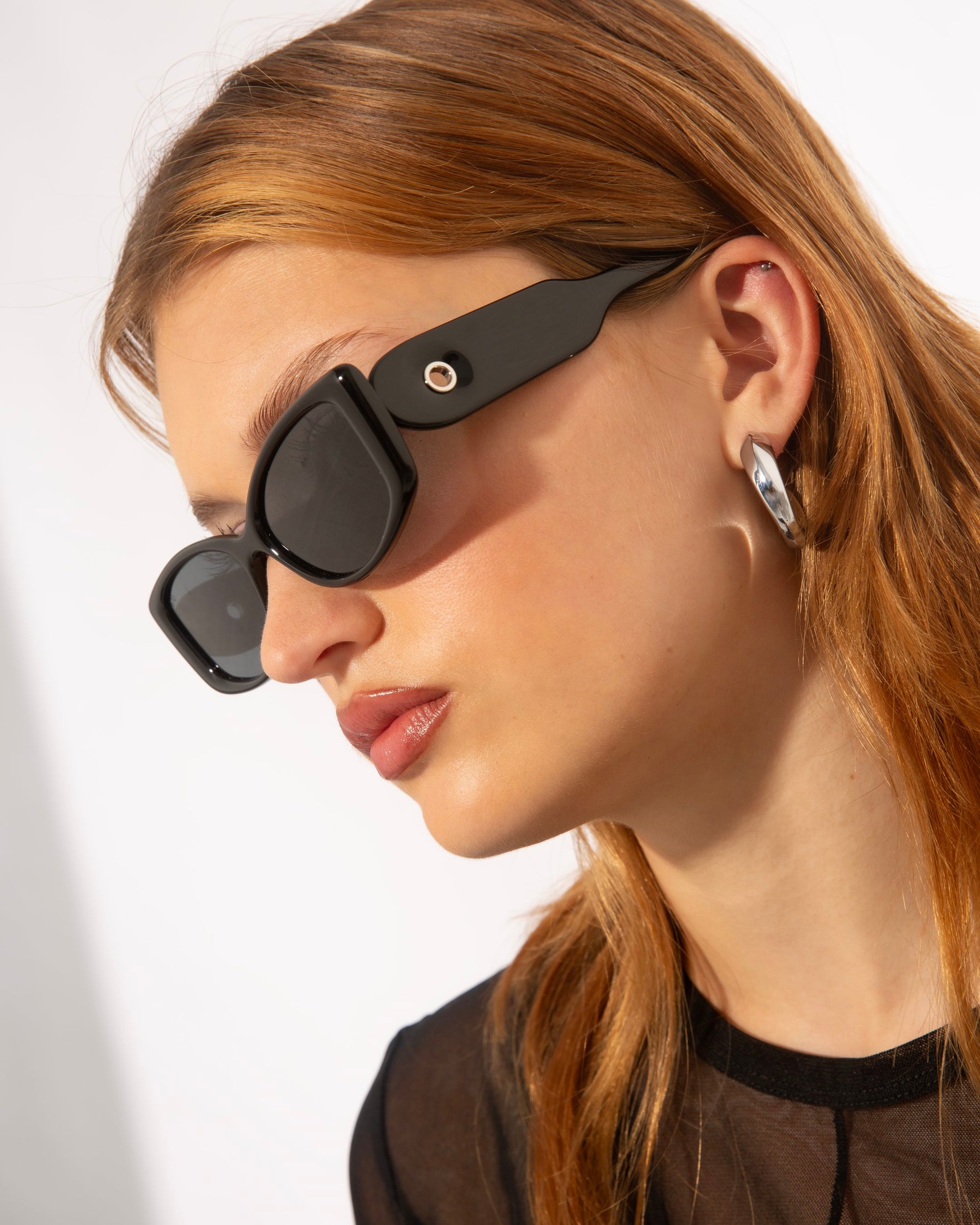 A person with light brown hair is wearing black For Art's Sake® Mene sunglasses and silver hoop earrings. They are dressed in a black top and are looking to the side, showcasing their chic Cat-Eye silhouette. The background is plain white.