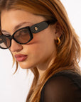 A person with long reddish hair wears black rectangular Mene sunglasses by For Art's Sake® and a black top. They have 18 karat gold hoop earrings and are looking to the side, set against a plain white background.