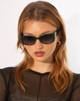 A woman with long, light brown hair wears angular, dark green For Art's Sake® Mene sunglasses with a subtle Cat-Eye silhouette and gold hoop earrings featuring 18 karat gold plating. She has a serious expression and is dressed in a black, sheer top, posing against a plain white background.