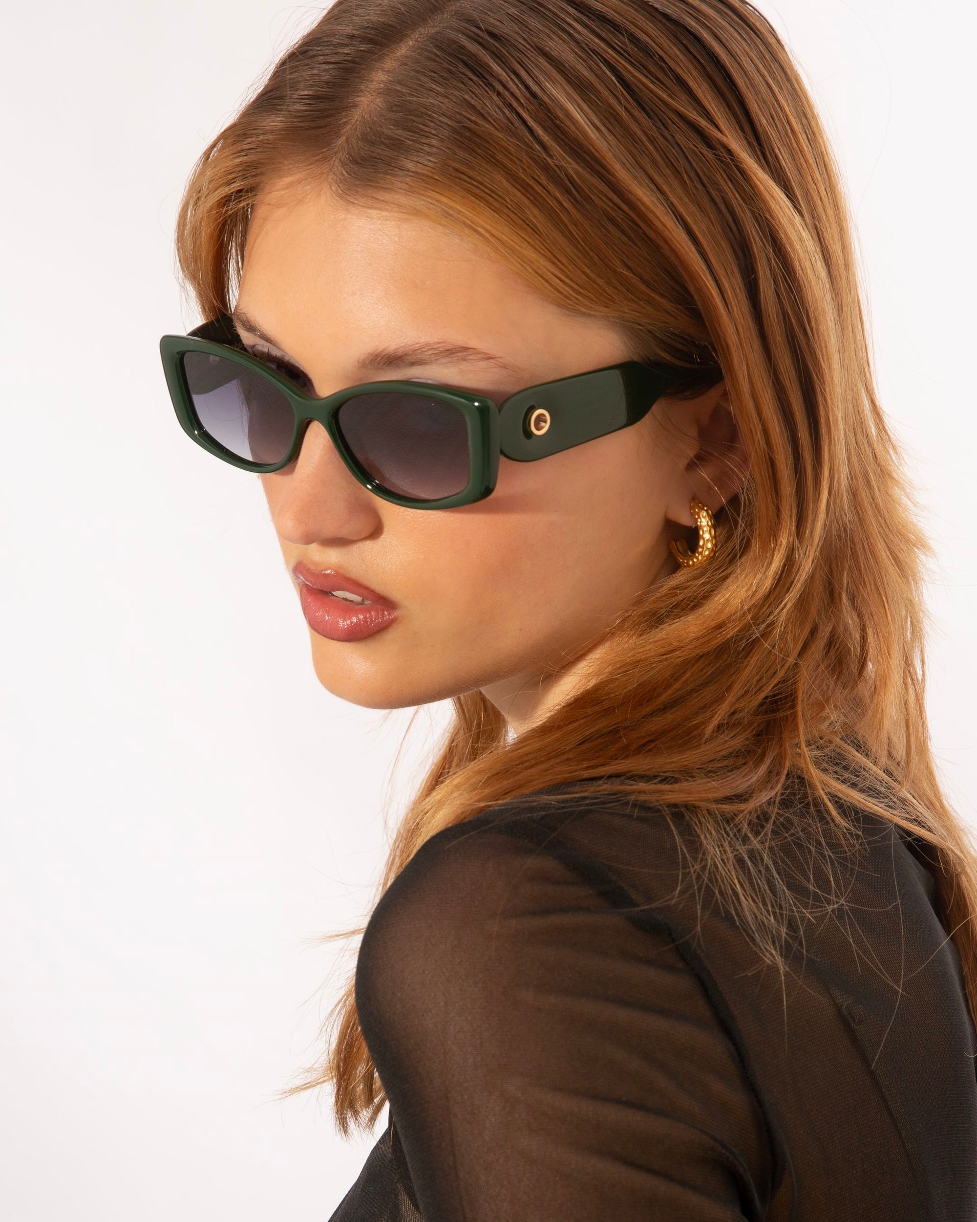 A woman with long light brown hair wearing dark green For Art&#39;s Sake® Mene sunglasses with a Cat-Eye silhouette and gold hoop earrings. She is dressed in a sheer black top and is looking to her left. The background is plain white.