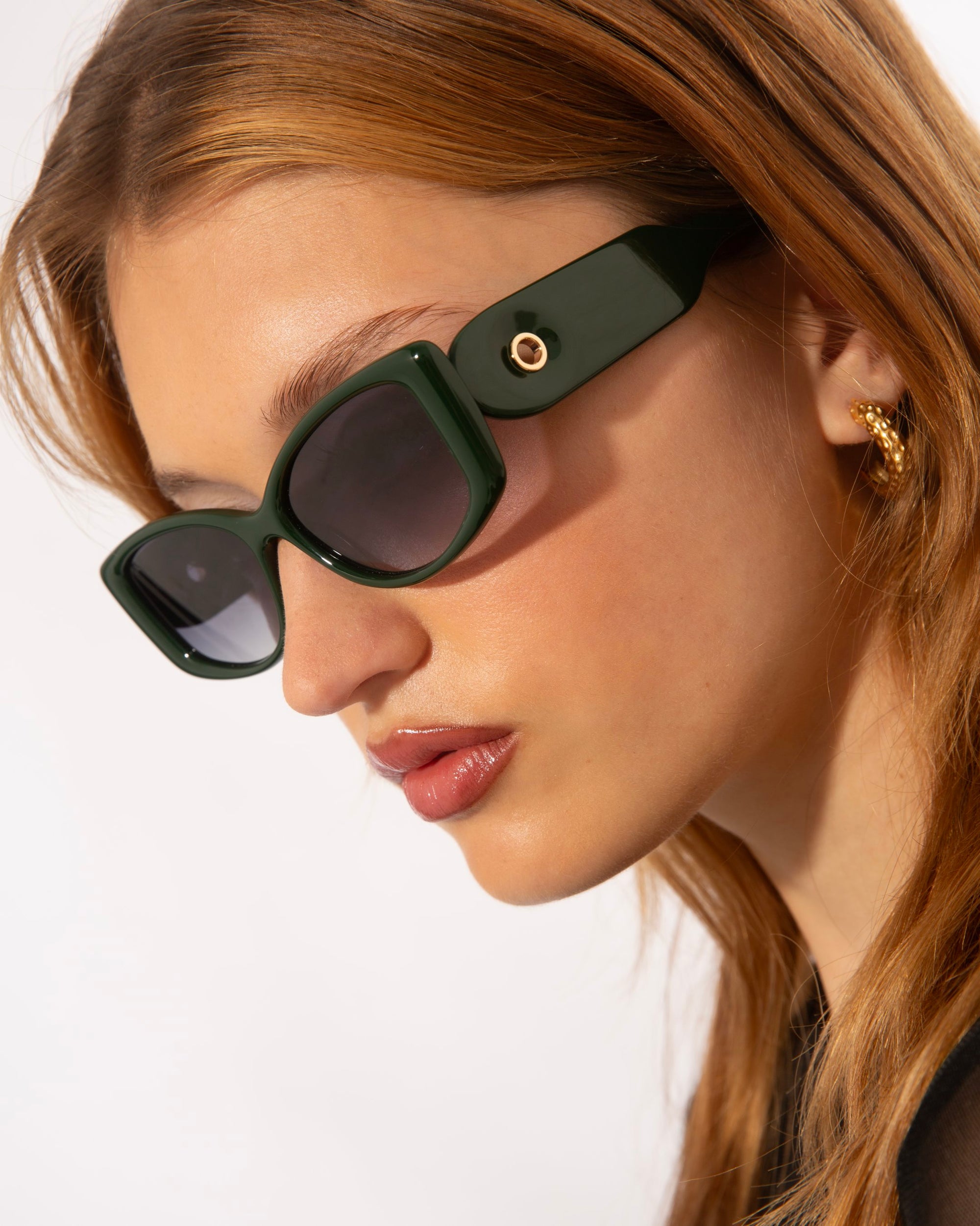 A person with long, auburn hair is wearing dark green, rectangular Mene sunglasses by For Art&#39;s Sake® and gold hoop earrings. The background is a plain white, and the person is looking slightly downwards, showcasing the side profile of their face.