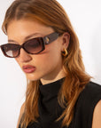 Young woman with shoulder-length light brown hair wearing Mene sunglasses by For Art's Sake® and gold hoop earrings. She is dressed in a high-neck black top and has a serious expression while looking down. The background is plain white.