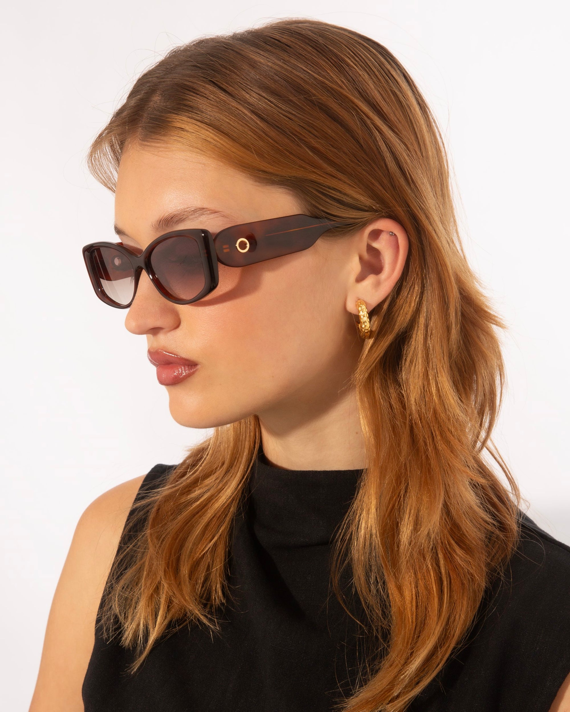 A person with long, reddish-brown hair, wearing 18 karat gold For Art&#39;s Sake® Mene sunglasses and gold hoop earrings, looks to the side. They are dressed in a sleeveless black top against a plain white background.