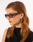 A person with long, reddish-brown hair, wearing 18 karat gold For Art's Sake® Mene sunglasses and gold hoop earrings, looks to the side. They are dressed in a sleeveless black top against a plain white background.