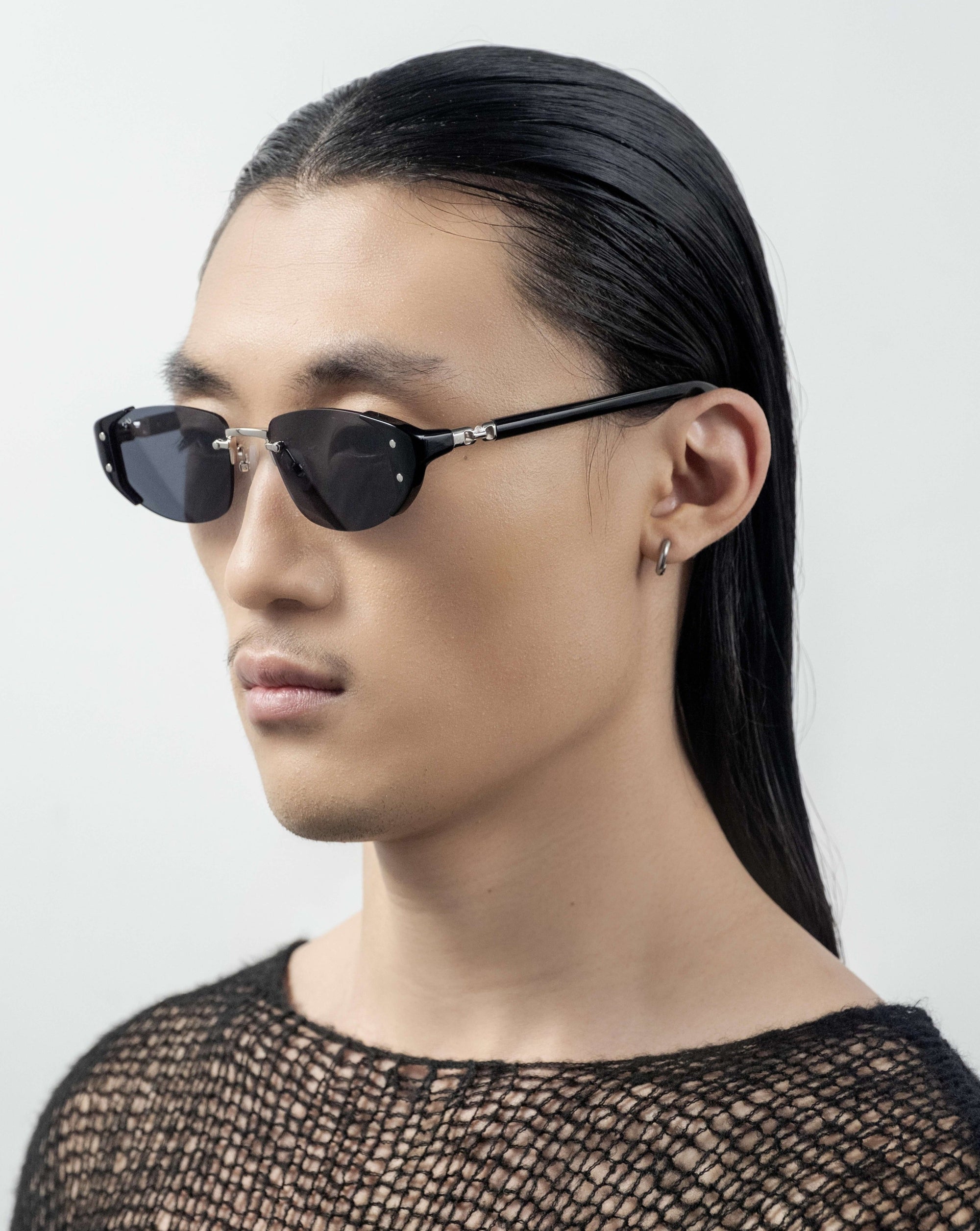 A person with long, sleek black hair and wearing For Art&#39;s Sake® Harbour sunglasses with a retro-inspired design looks to the side. They have a small hoop earring in one ear and are dressed in a textured, black mesh top. The background is plain white.