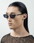 A person with long, sleek black hair and wearing For Art's Sake® Harbour sunglasses with a retro-inspired design looks to the side. They have a small hoop earring in one ear and are dressed in a textured, black mesh top. The background is plain white.