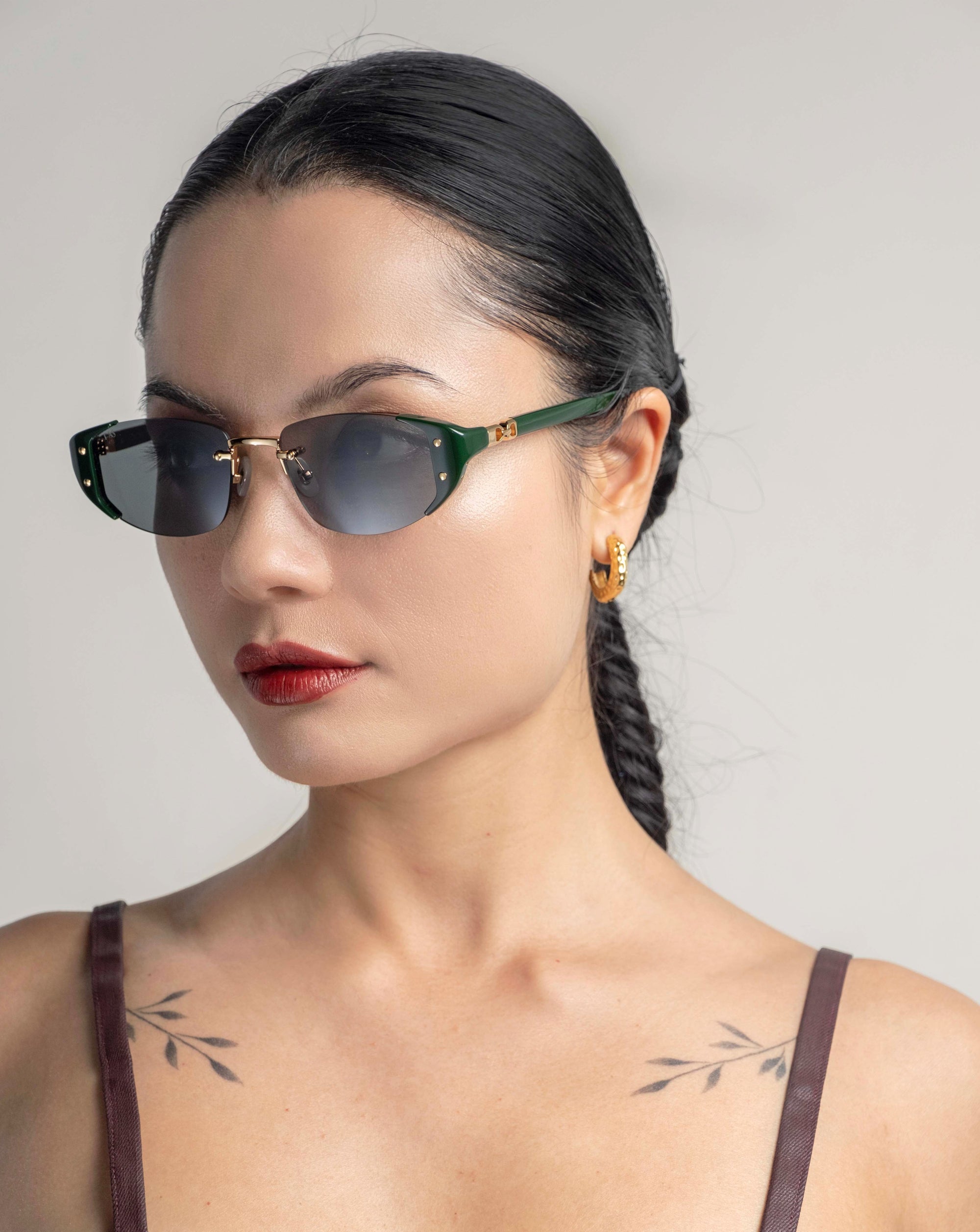 A woman with dark hair tied in a braid is wearing Harbour sunglasses by For Art&#39;s Sake® and a brown strappy top. She accessorizes with 18 karat gold-plated hoop earrings and has small branch-like tattoos on her chest. The background is plain and light-colored, adding to the elegant, retro-inspired design of her look.