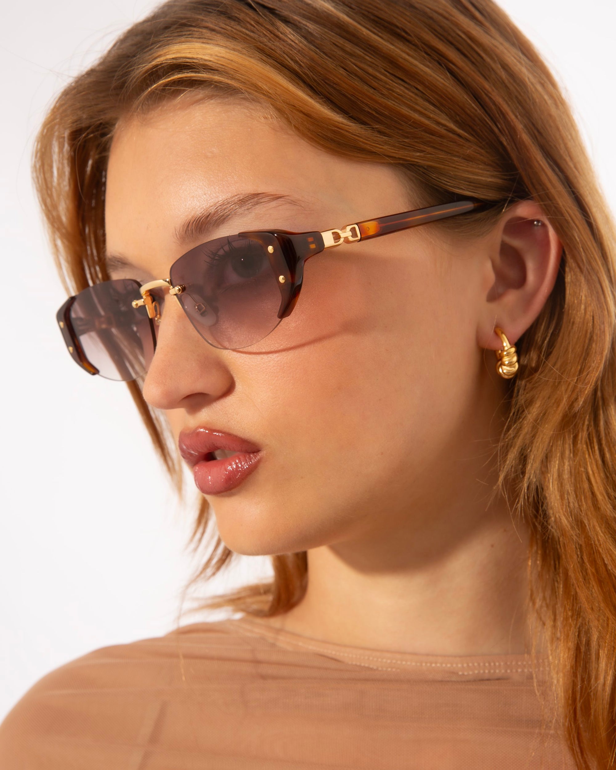 A woman with long, light brown hair is wearing stylish For Art's Sake® Harbour sunglasses featuring retro-inspired design with gold accents and small hoop earrings. She is dressed in a sheer, nude-colored top. The background is plain white, emphasizing her fashion accessories.