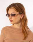 A woman with light brown hair is wearing retro-inspired Harbour sunglasses by For Art's Sake® and small gold earrings with 18 karat gold plating. She has a neutral expression and is looking to her left, dressed in a sheer, light brown top against a plain white background.