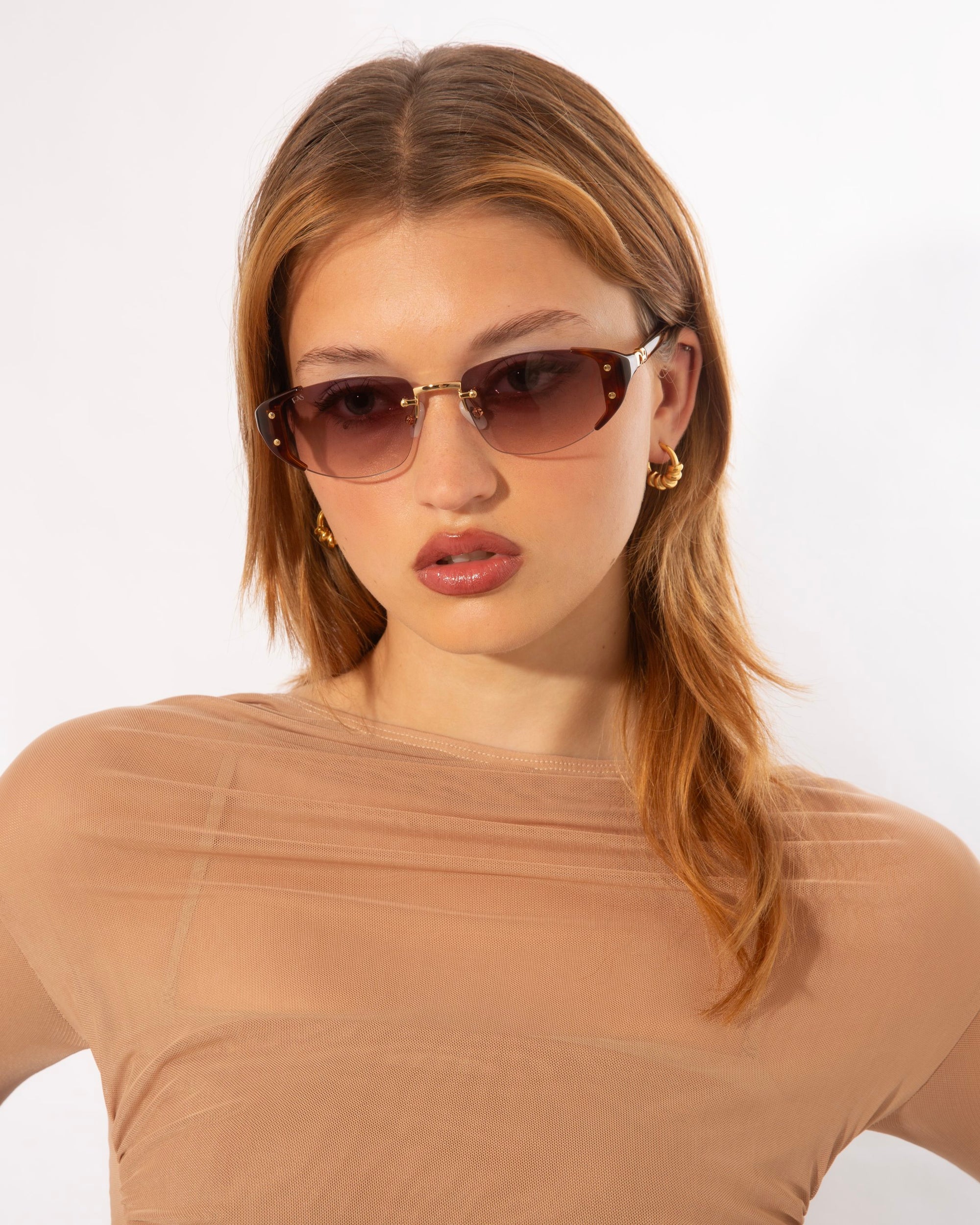 A person with long, light brown hair wearing stylish Harbour sunglasses and gold hoop earrings is pictured from the waist up. They are dressed in a sheer, beige top against a plain white background. The retro-inspired design of the For Art&#39;s Sake® Harbour sunglasses adds a modern twist to their look.