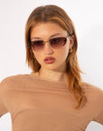 A person with long, light brown hair wearing stylish Harbour sunglasses and gold hoop earrings is pictured from the waist up. They are dressed in a sheer, beige top against a plain white background. The retro-inspired design of the For Art's Sake® Harbour sunglasses adds a modern twist to their look.