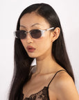 A woman with long, straight black hair is wearing For Art's Sake® Harbour sunglasses featuring light gray frames and jade-stone nose pads. She has a small earring in one ear and is dressed in a spaghetti strap top with lace details. She looks directly ahead against a plain white background.