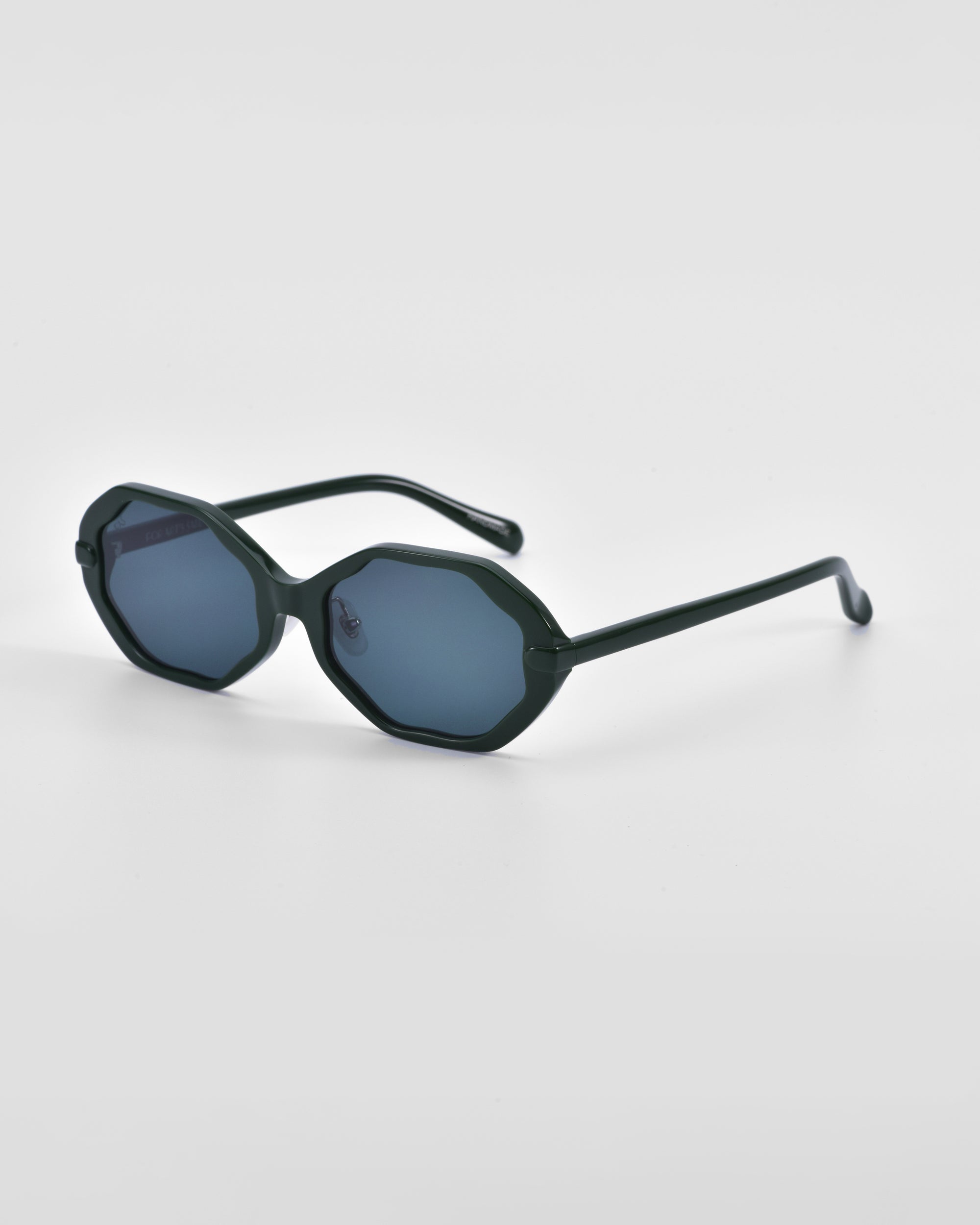 A pair of For Art&#39;s Sake® Lotus sunglasses featuring a dark green angular silhouette and dark tinted lenses, complemented by thin matching green arms and jade-stone nose pads, placed on a plain white background.