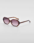 A pair of For Art's Sake® Lotus sunglasses with tortoiseshell frames and round pink-tinted lenses is placed against a white background. The frame, featuring an angular silhouette, has a distinct, slightly geometric design and slender arms.