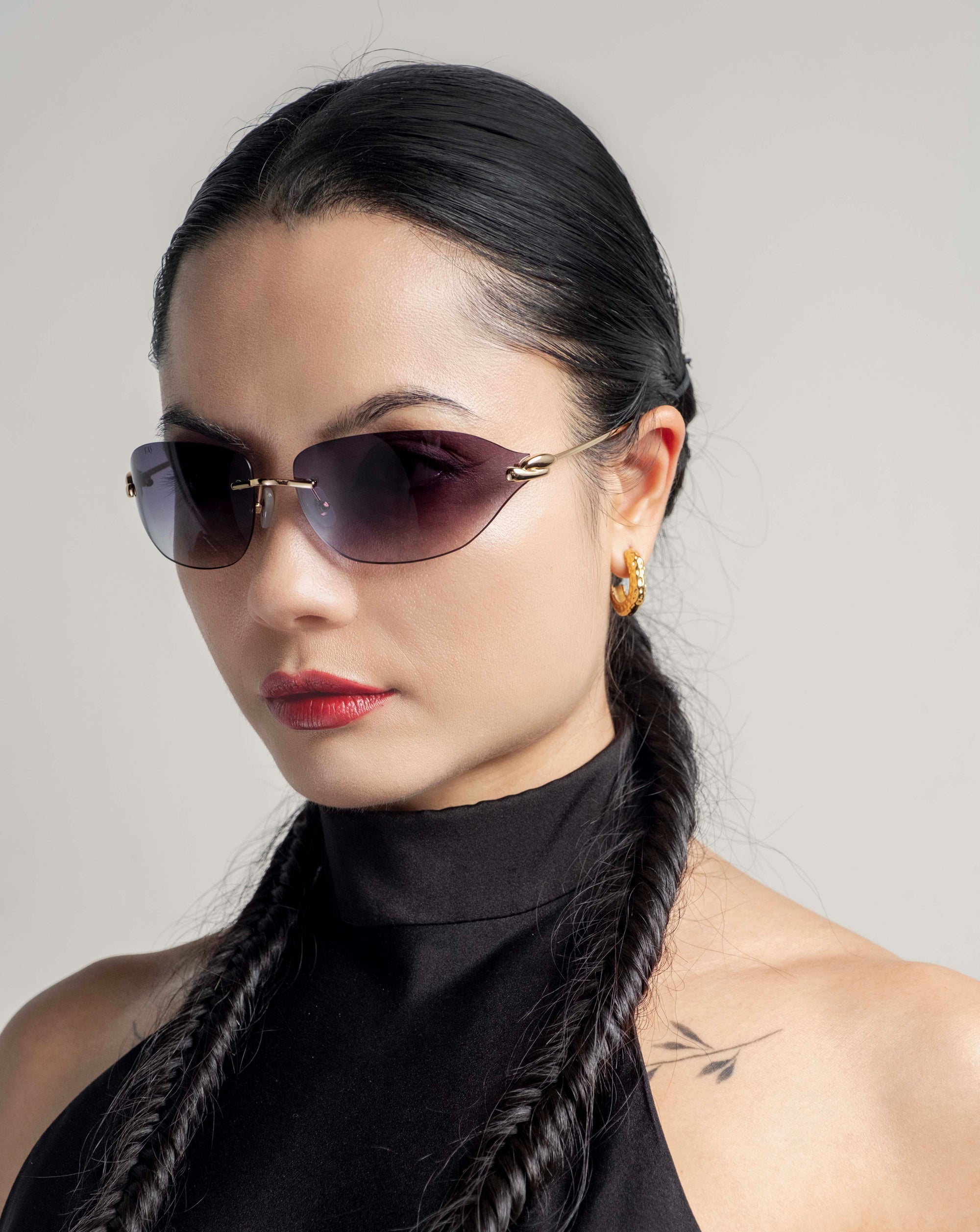 A woman with long, dark braided hair and 18-karat gold hoop earrings wears For Art&#39;s Sake® Serpent I sunglasses featuring a bold frameless wrap lens design. She has a tattoo near her shoulder and is looking confidently into the camera against a neutral background in her sleeveless black top.
