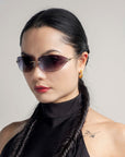 A woman with long, dark braided hair and 18-karat gold hoop earrings wears For Art's Sake® Serpent I sunglasses featuring a bold frameless wrap lens design. She has a tattoo near her shoulder and is looking confidently into the camera against a neutral background in her sleeveless black top.