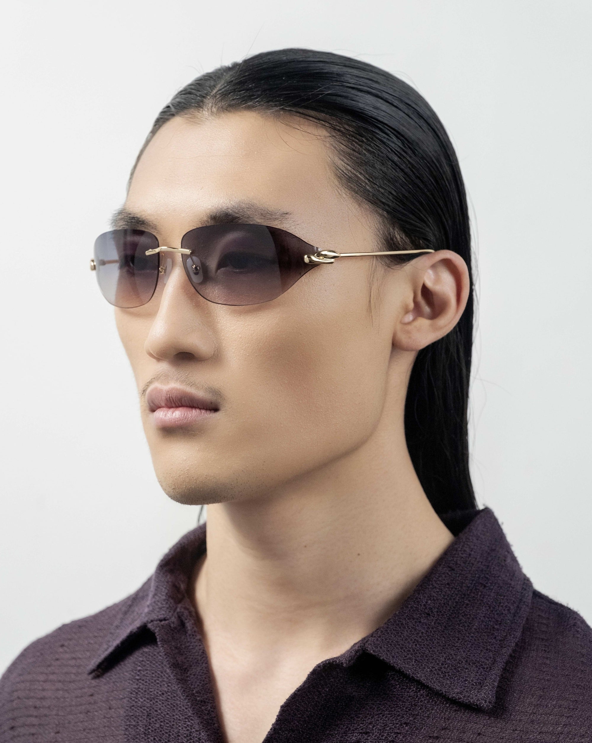 A person with long, sleek dark hair is seen wearing For Art&#39;s Sake® Serpent I frameless wrap lens oval-shaped sunglasses with golden temple arms. They are dressed in a dark, textured shirt and facing slightly left against a plain, light background.