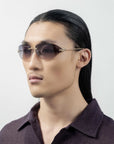 A person with long, sleek dark hair is seen wearing For Art's Sake® Serpent I frameless wrap lens oval-shaped sunglasses with golden temple arms. They are dressed in a dark, textured shirt and facing slightly left against a plain, light background.