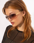 A woman with light brown, shoulder-length hair wears oversized sunglasses with thin golden frames and For Art's Sake® Serpent I detailing. She is also wearing a black, sheer top and textured golden hoop earrings. The background is plain white.