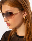 A close-up side profile of a person with long light brown hair, wearing Serpent I by For Art's Sake® and gold hoop earrings. The person is dressed in a sheer black top, and the background is plain white.