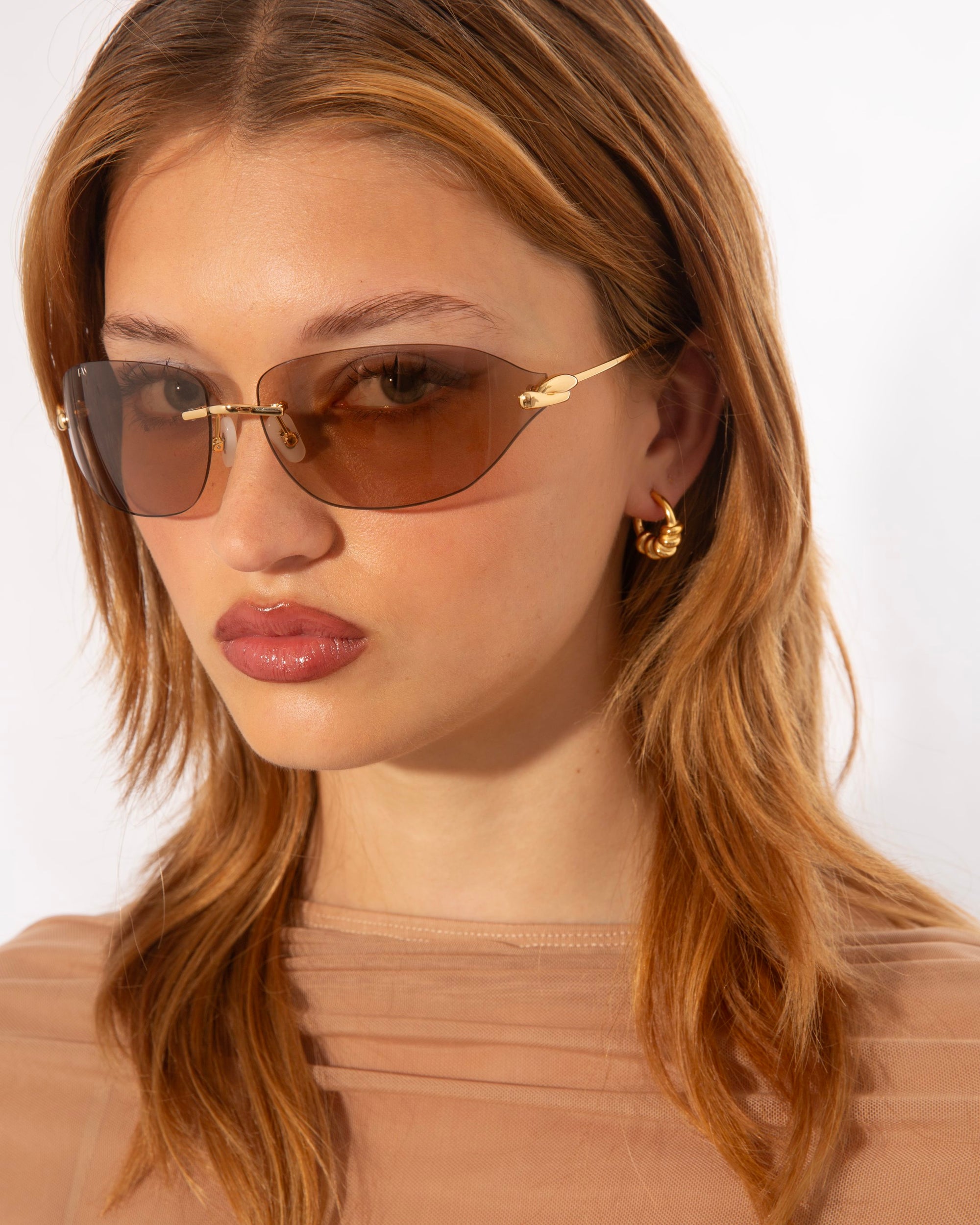 A woman with shoulder-length light brown hair is wearing stylish, For Art's Sake® Serpent I sunglasses and small gold hoop earrings. She is dressed in a light brown sheer top and is posing against a plain white background.