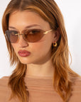 A woman with shoulder-length light brown hair is wearing stylish, For Art's Sake® Serpent I sunglasses and small gold hoop earrings. She is dressed in a light brown sheer top and is posing against a plain white background.