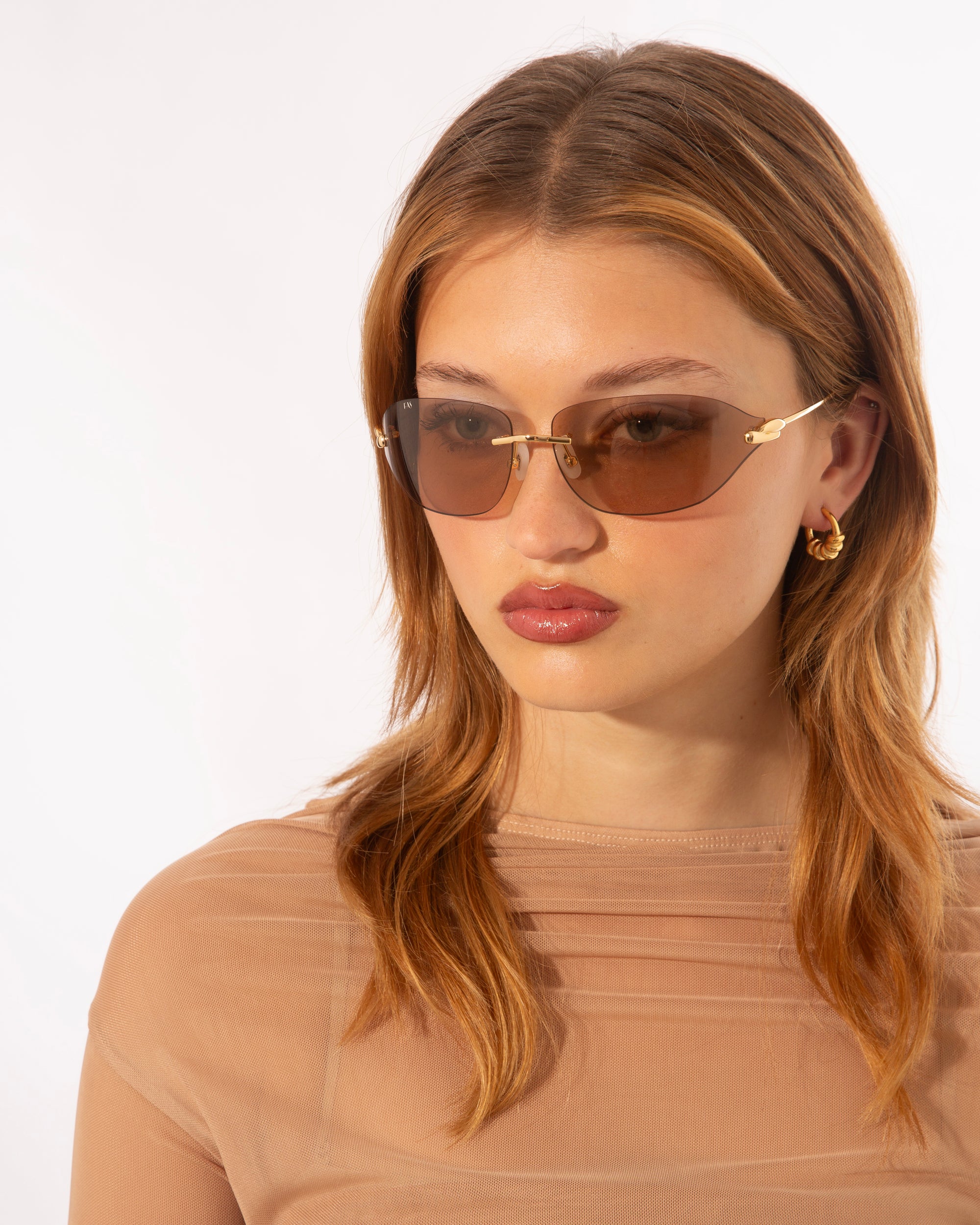 A person with long brown hair wearing For Art&#39;s Sake® Serpent I sunglasses and gold hoop earrings. They are dressed in a translucent beige top and look slightly to the side against a plain white background.