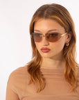 A person with long brown hair wearing For Art's Sake® Serpent I sunglasses and gold hoop earrings. They are dressed in a translucent beige top and look slightly to the side against a plain white background.