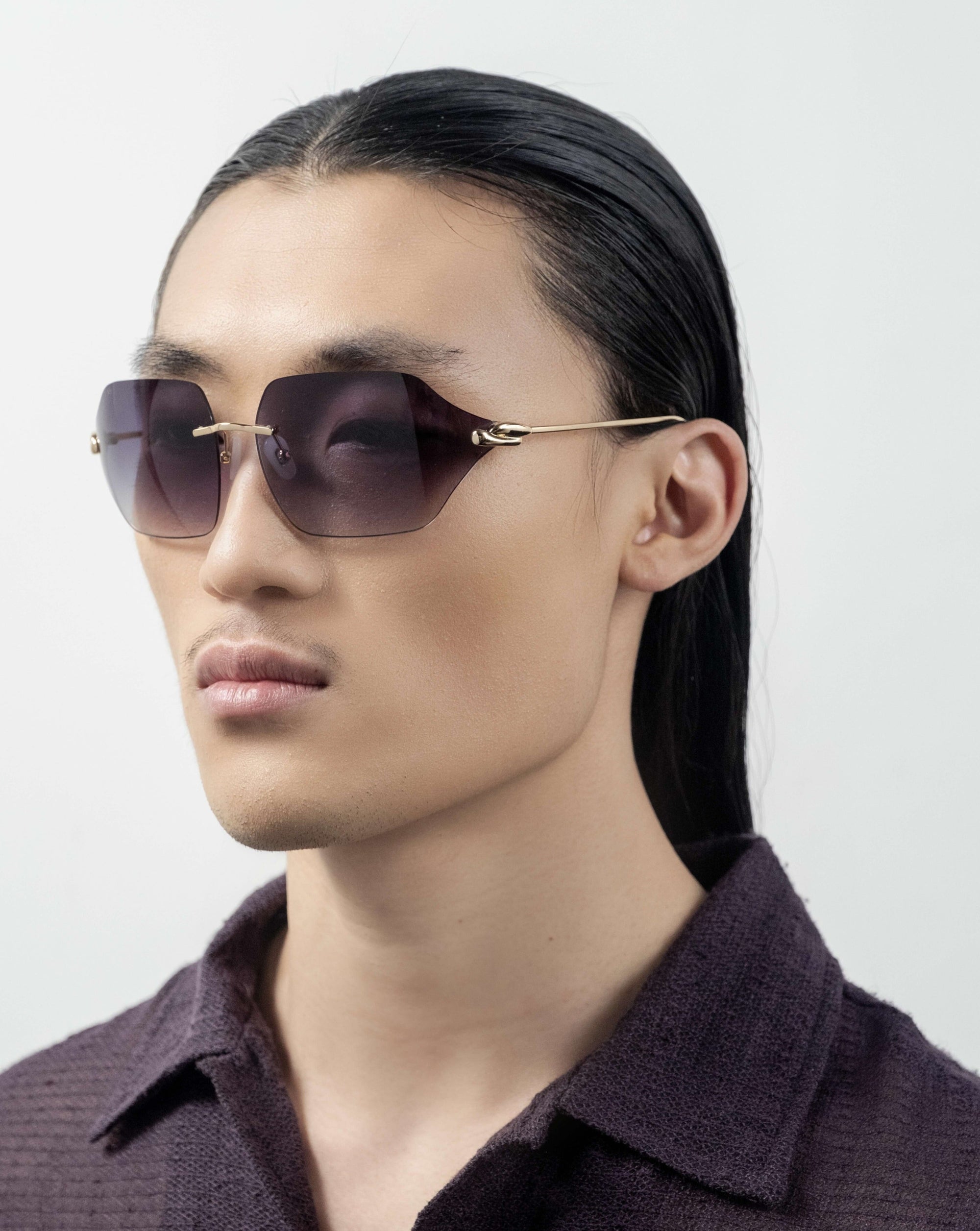 A person with long, dark hair wearing For Art's Sake® Serpent II sunglasses featuring a frameless wrap lens design and a dark textured shirt stares confidently ahead. The background is plain and white, emphasizing their serious expression and modern, stylish appearance.