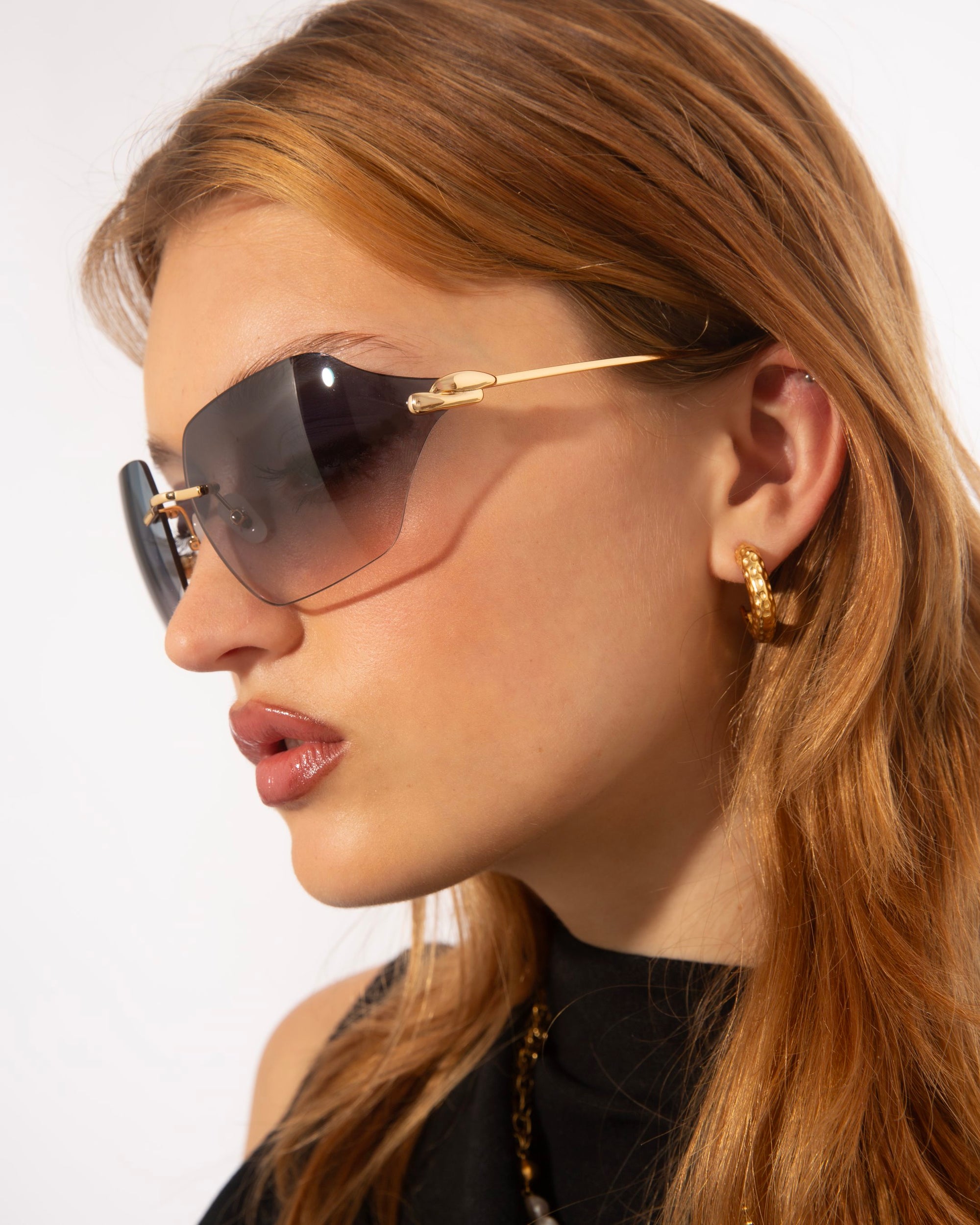 A woman with long, reddish-brown hair is pictured in profile wearing For Art&#39;s Sake® Serpent II sunglasses with jade-stone nose pads and gold hoop earrings. She is dressed in a black top, and the background is plain white. The image focuses on her head and shoulders, highlighting her accessories.