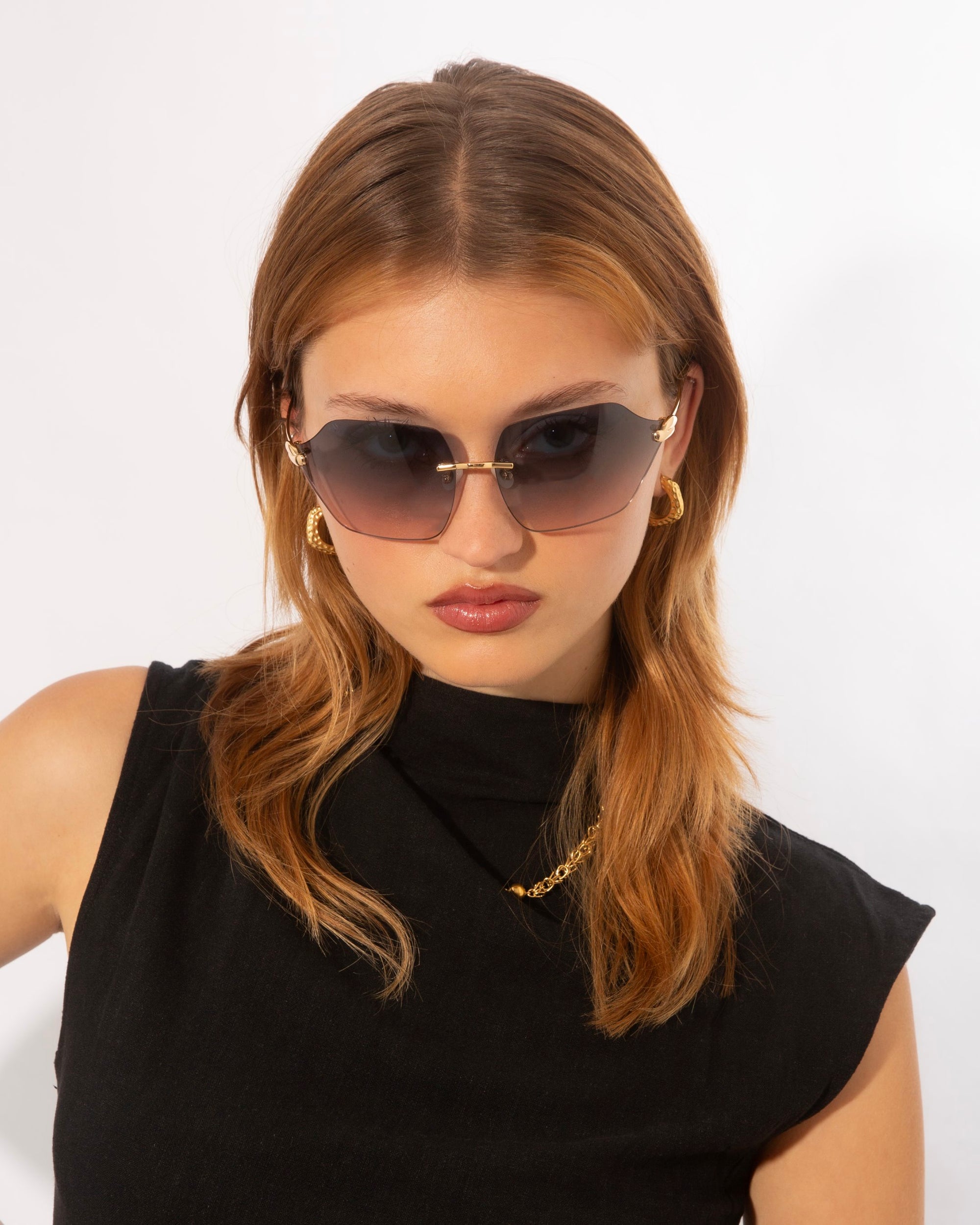 A person with light brown hair wearing large, dark sunglasses (Serpent II by For Art's Sake®) with frameless wrap lenses and gold earrings looks directly at the camera. They are dressed in a sleeveless black top against a plain white background.