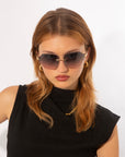 A person with light brown hair wearing large, dark sunglasses (Serpent II by For Art's Sake®) with frameless wrap lenses and gold earrings looks directly at the camera. They are dressed in a sleeveless black top against a plain white background.