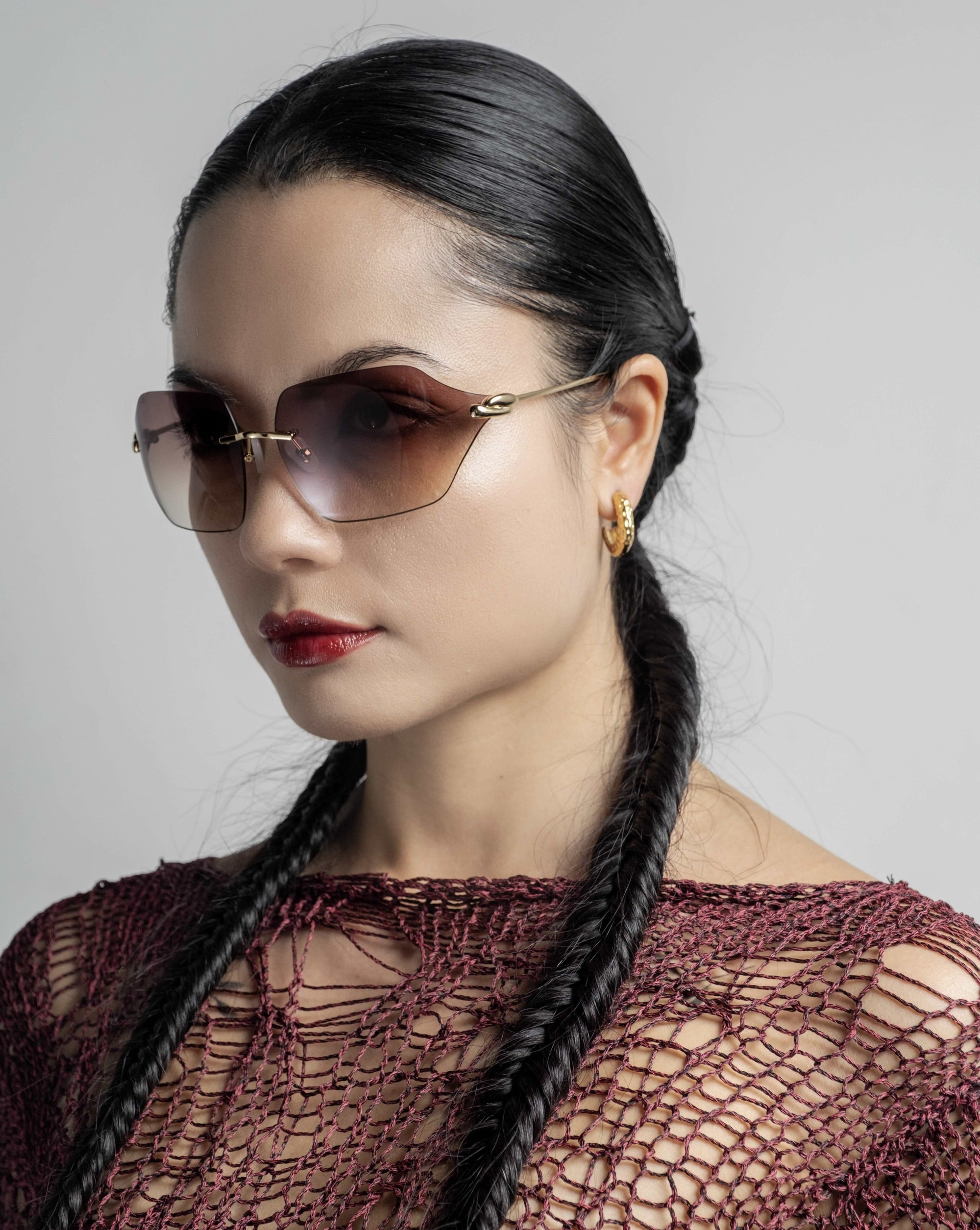 A woman with dark hair styled in two long braids wears For Art&#39;s Sake® Serpent II sunglasses, golden hoop earrings, and a burgundy crochet top. She has a neutral facial expression and is set against a plain, light gray background.