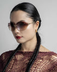 A woman with dark hair styled in two long braids wears For Art's Sake® Serpent II sunglasses, golden hoop earrings, and a burgundy crochet top. She has a neutral facial expression and is set against a plain, light gray background.